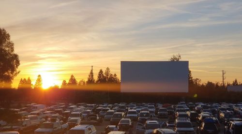 A sunset over a crowded drive-in movie theater with a large blank screen and parked cars.