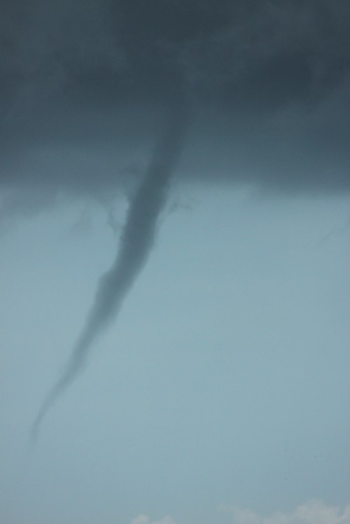 A slender tornado touches down from dark storm clouds against a light blue sky.