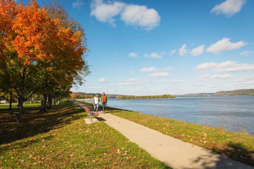Two people walk along a scenic path by a river, surrounded by colorful autumn trees and a clear blue sky.