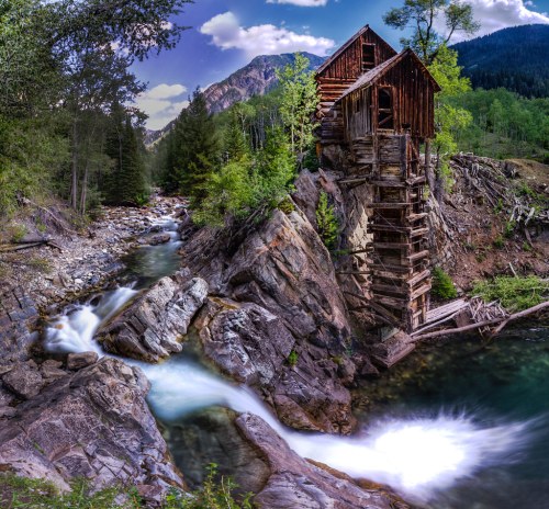 A rustic wooden structure perched on a rocky cliff beside a flowing river, surrounded by lush green trees and mountains.