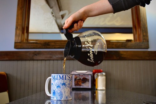 A hand pours coffee from a glass carafe into a blue mug on a table in a cozy diner setting.