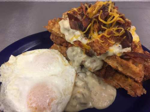 A plate of crispy waffles topped with cheese, bacon, and gravy, served alongside two sunny-side-up eggs.