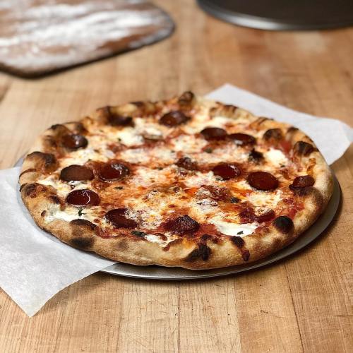A freshly baked pepperoni pizza on a metal tray, resting on a wooden table with flour dust in the background.