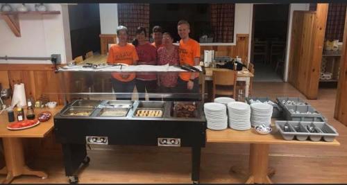 A group of five people stands behind a buffet table in a dining area, with food and dishes arranged around them.