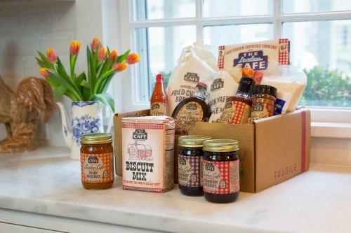 A box of assorted food items, including biscuit mix and preserves, sits on a kitchen counter beside a vase of tulips.