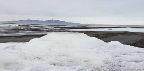 A frozen landscape with ice formations in the foreground and distant mountains under a cloudy sky.