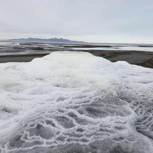 A frozen landscape with textured ice formations and a calm sea in the background, under a cloudy sky.