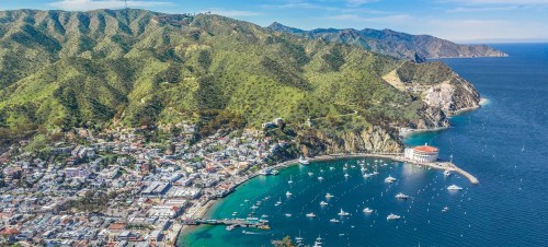 Aerial view of a coastal town surrounded by mountains, featuring a harbor with boats and a distinctive round building.