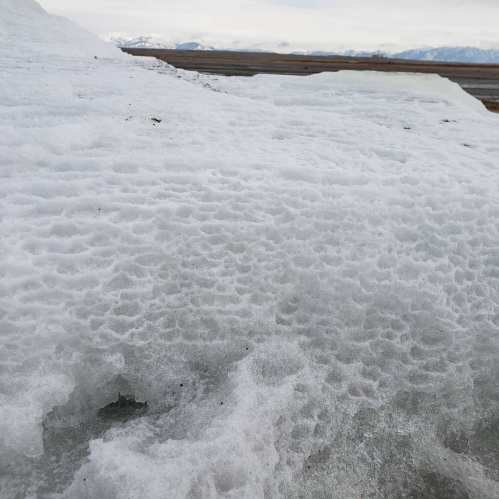 Close-up of textured ice with a snowy landscape and mountains in the background under a cloudy sky.