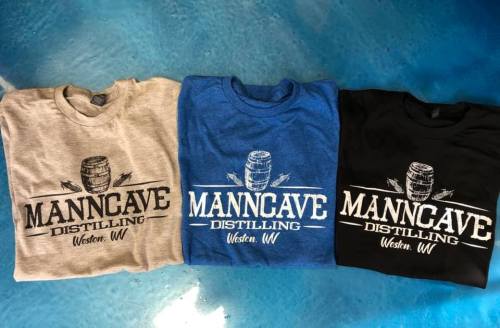 Three t-shirts in gray, blue, and black featuring "MannCave Distilling" logo with a barrel design.