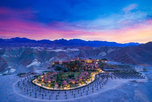 Aerial view of a resort surrounded by mountains at sunset, with vibrant skies and illuminated pathways.