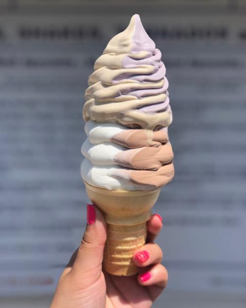 A hand holding a multi-flavored ice cream cone with swirls of purple, brown, and white against a blurred background.