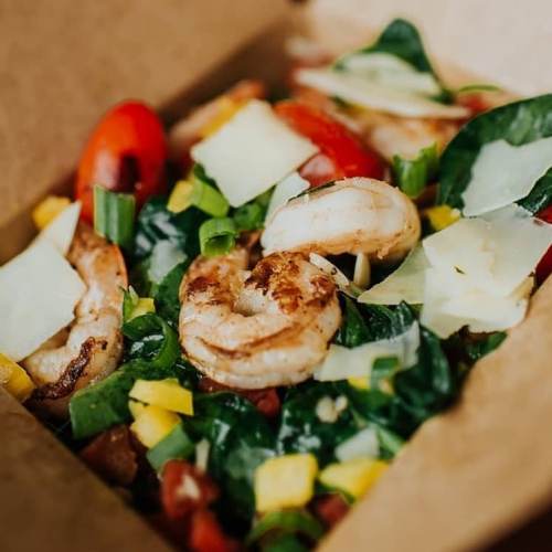 A colorful salad with shrimp, cherry tomatoes, mango, greens, and cheese, served in a brown takeout box.
