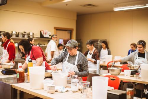 A busy cooking class with participants in aprons, measuring ingredients and preparing food at various workstations.