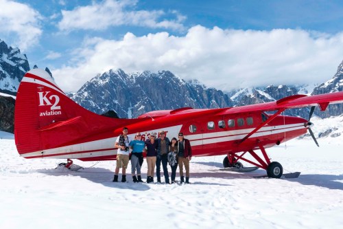 A group of five people stands in front of a red plane on a snowy landscape with mountains in the background.
