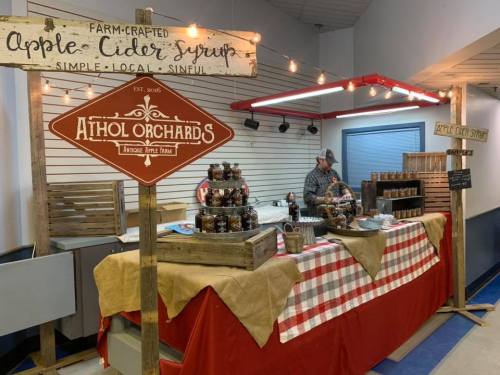 A market stall featuring apple cider syrup, jars, and a vendor behind a checkered tablecloth with signs for Athol Orchards.