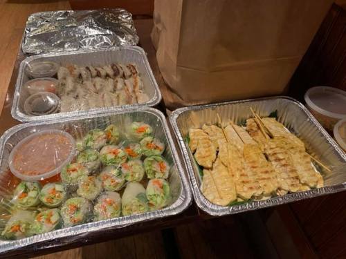 A table with trays of food, including spring rolls, grilled chicken skewers, and dipping sauces.