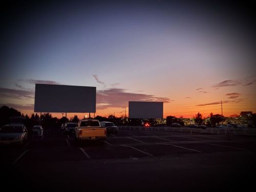 Two blank billboards stand in a parking lot at sunset, with colorful clouds and parked cars in the foreground.