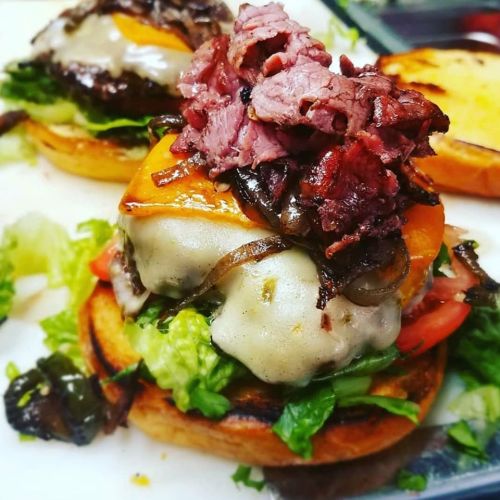 A close-up of a delicious burger topped with melted cheese, lettuce, tomato, grilled onions, and sliced meat on a toasted bun.