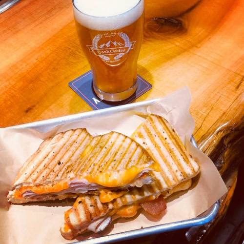 A grilled sandwich with melted cheese and meats, served with a pint of beer on a wooden table.