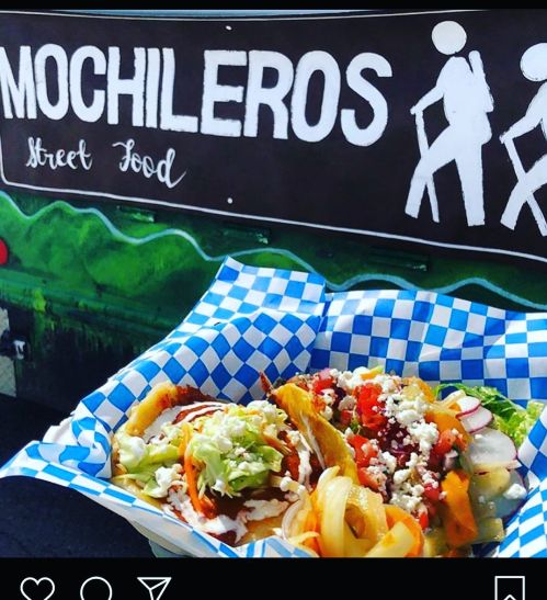 Two tacos with colorful toppings served in a blue and white checkered basket, with a food truck in the background.