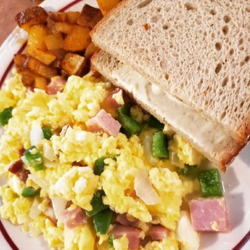 A plate with scrambled eggs mixed with ham and green peppers, a slice of bread, and golden-brown diced potatoes.