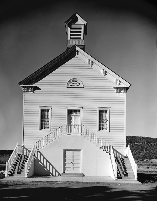 A historic white schoolhouse with a bell tower, featuring two sets of stairs and large windows.