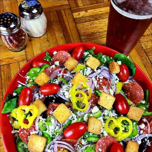 A vibrant salad with greens, tomatoes, olives, onions, croutons, and pepper rings, alongside a glass of dark beer.