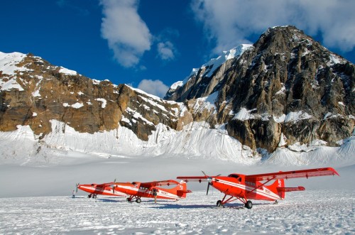 Three red airplanes parked on a snowy landscape with towering mountains and a blue sky in the background.