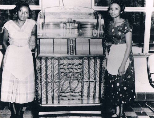 Two women in vintage dresses stand beside a classic jukebox in a retro diner setting.