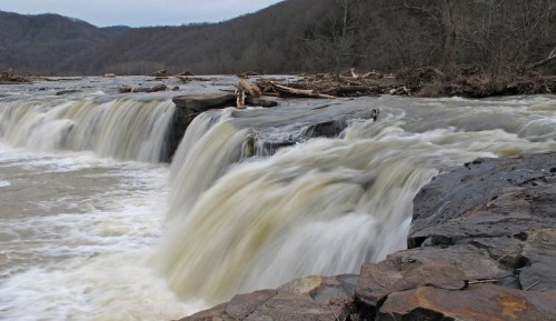 A cascading waterfall flows over rocky ledges, surrounded by trees and mountains in the background.