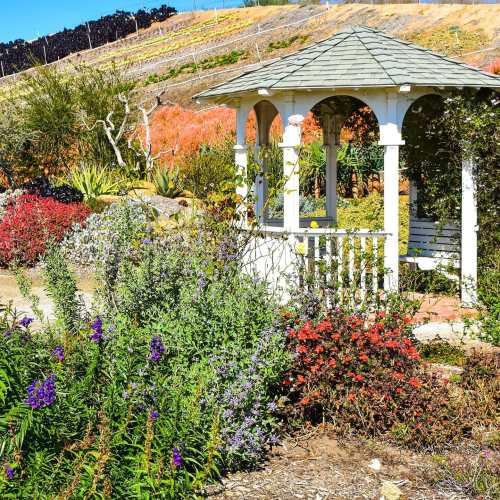 A white gazebo surrounded by colorful flowers and lush greenery in a vibrant garden setting.