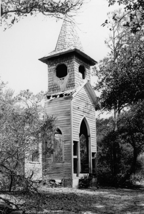 A dilapidated wooden church with a steeple, surrounded by overgrown trees and vegetation.