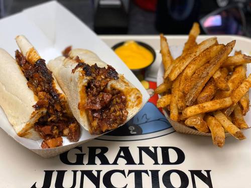 Two loaded hot dogs with toppings, served with a side of seasoned fries and a small cup of mustard.
