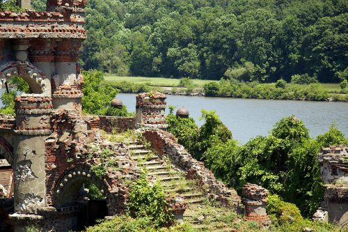 Ruins of a brick structure overgrown with greenery, beside a calm river and lush trees in the background.