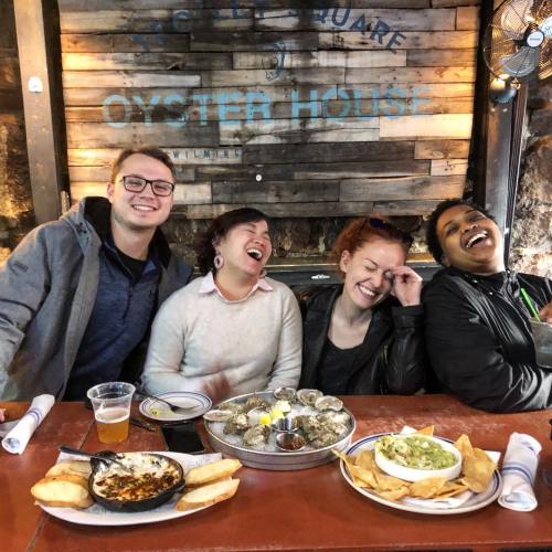 Four friends laughing at a table with seafood dishes, including oysters and a dip, in a cozy restaurant setting.