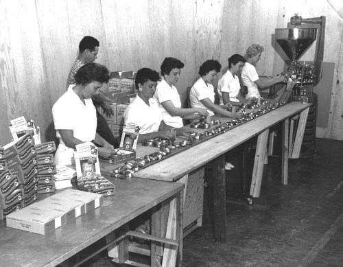 Black and white photo of women working at a long table, assembling products in a warehouse setting.