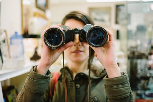 A person holds binoculars up to their eyes, looking through them in a vintage shop.