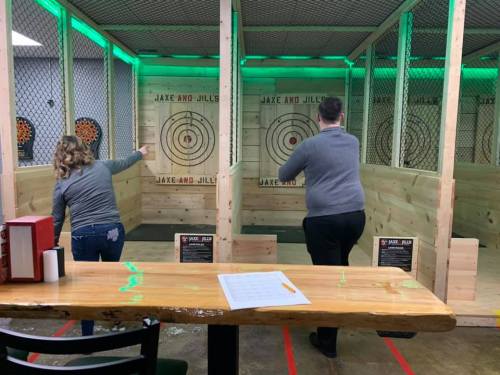 Two people throwing axes at targets in a well-lit indoor axe-throwing venue with wooden barriers.