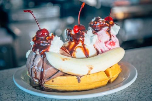 A banana split with three scoops of ice cream, topped with chocolate sauce, whipped cream, and cherries, served on a waffle.