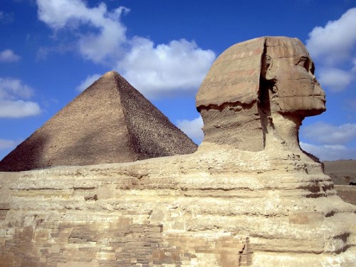 The Great Sphinx of Giza with the Pyramid of Khufu in the background under a blue sky with clouds.