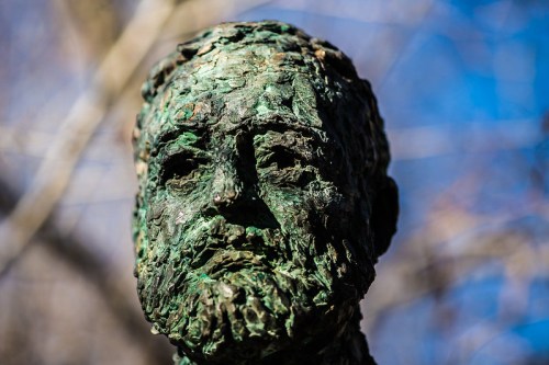 Close-up of a weathered bronze statue of a bearded man, with a textured surface and a blurred natural background.