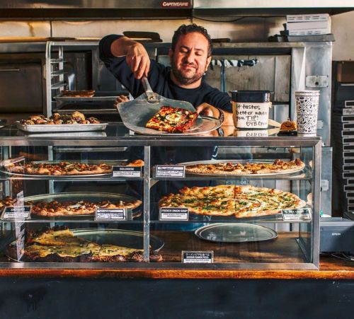 A chef serving pizza from a display case in a bustling pizzeria, showcasing various pizza options.