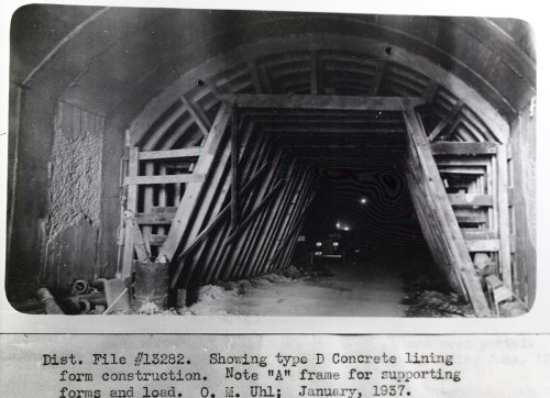 Black and white photo of a concrete tunnel under construction, featuring wooden supports and a vehicle in the background.