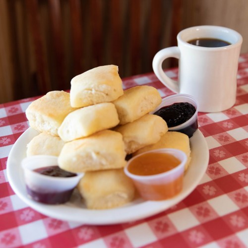 A plate of fluffy biscuits with three small cups of jam and a cup of coffee on a checkered tablecloth.