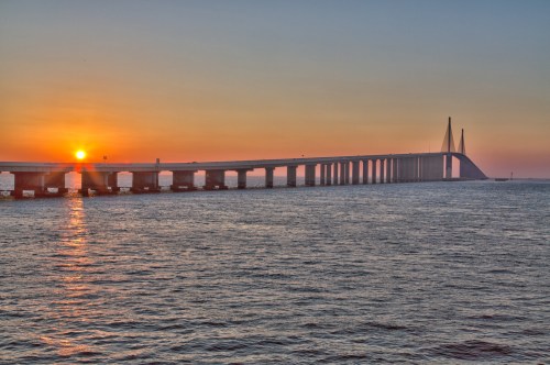 A sunset over a long bridge extending across calm waters, with a clear sky and soft colors.