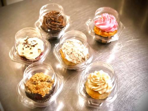 A variety of six cupcakes in clear plastic containers, featuring different frosting styles and toppings.