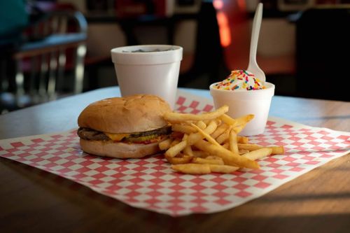 A cheeseburger with fries and a cup of ice cream with sprinkles on a checkered paper plate in a diner setting.