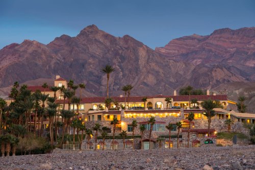 A scenic view of a resort nestled against mountains, illuminated at dusk with palm trees in the foreground.