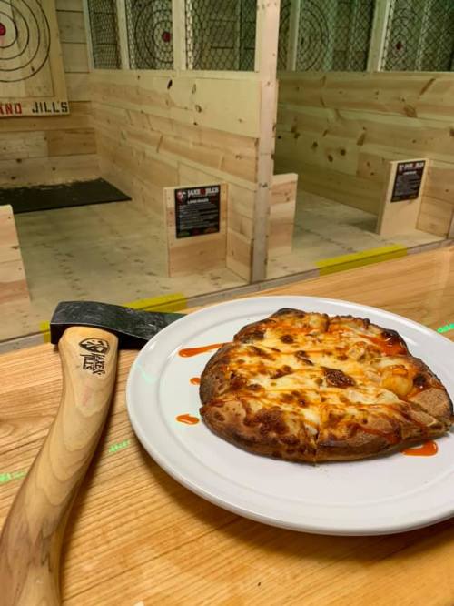 A plate of pizza with sauce sits on a wooden table next to an axe, with an axe-throwing range in the background.